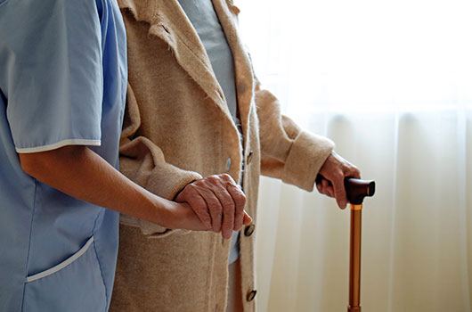 Auf dem Bild siehst du eine Seniorin, die am Stock geht und von einer Pflegerin dabei gestützt wird.