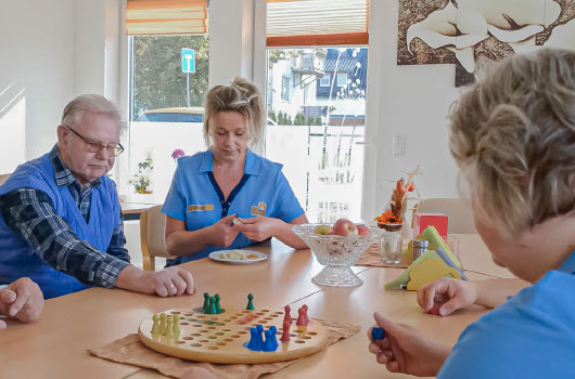 Auf dem Bild siehst du eine Pflegerin Äpfel schneidend mit Seniorinnen und Senioren beim gemeinsamen Mensch-ärgere-dich-nicht-Spiel.